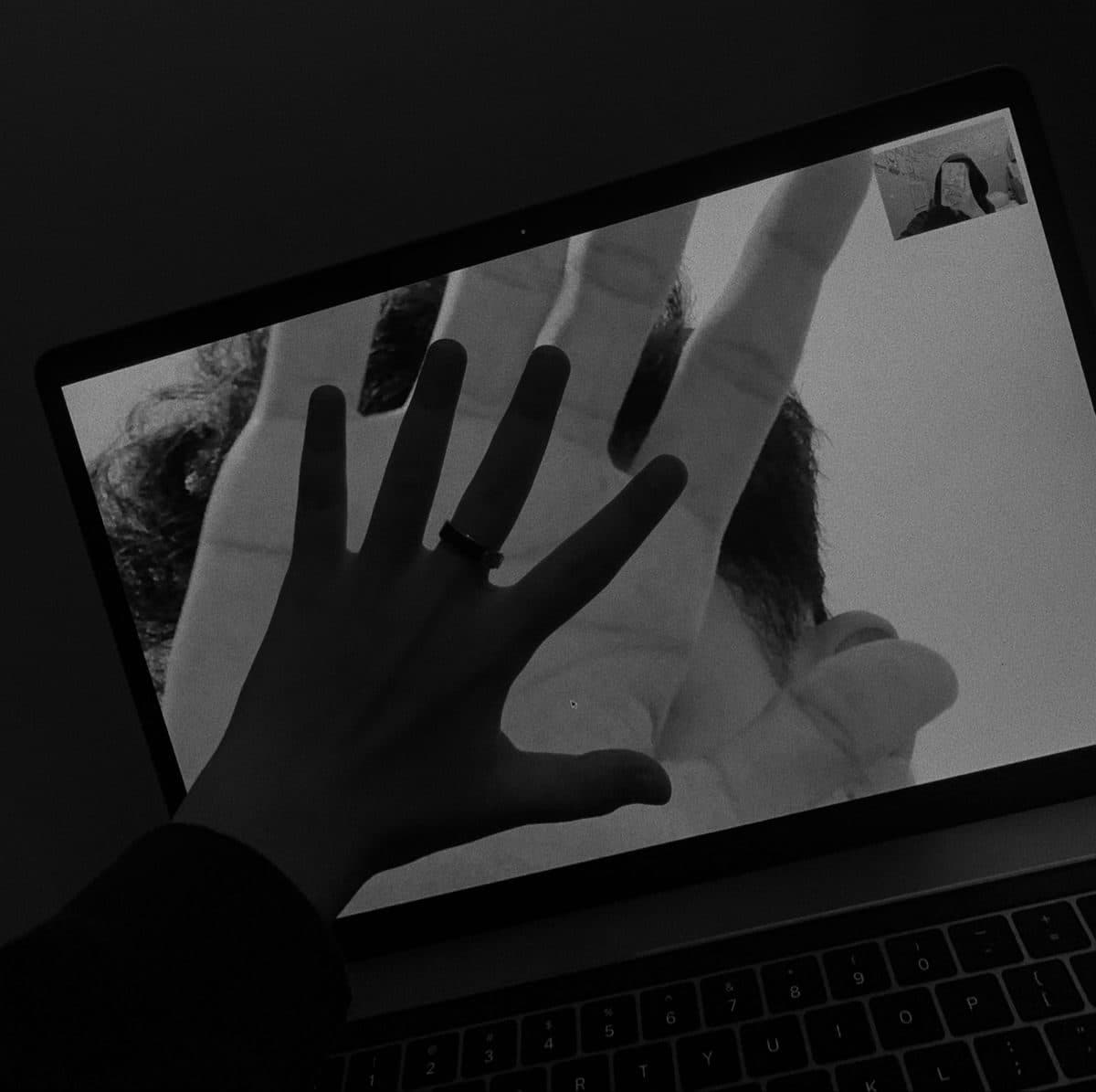A hand pressed to the laptop screen matching a partner's hand on a video call