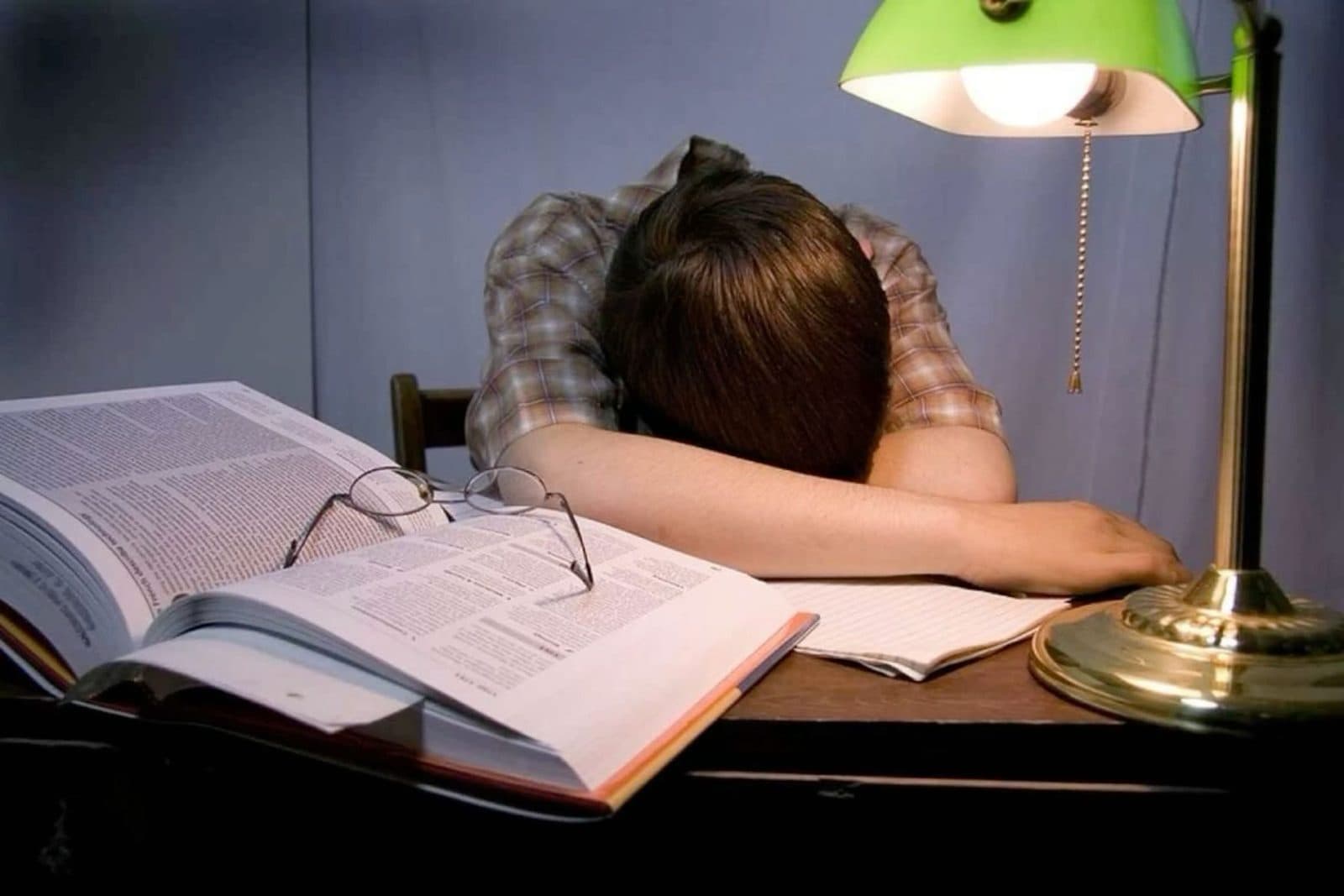 A tired student asleep on an open book under a desk lamp